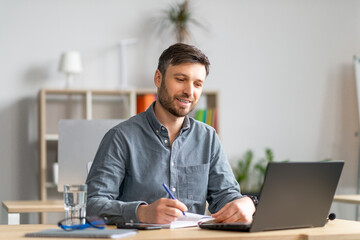 Poster - Happy mature entrepreneur man taking notes and using laptop, sitting at his workplace in office, copy space