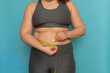 © Юля Бурмистрова - Unrecognizable fat plump overweight woman standing in grey sports bra, leggings, showing excess naked belly, trying to measure waist with tape on blue background. Dieting, unhealthy food, obesity.