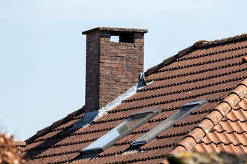 A portrait of a chimney on a roof of a house. The roof has ceramic tiles and two skylight windows in it which are closed.