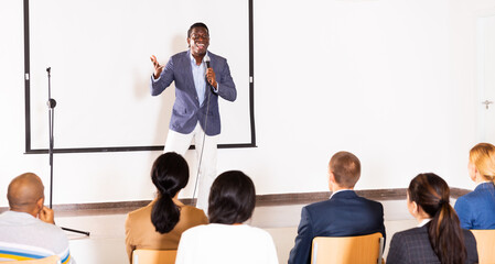 Wall Mural - Emotional aframerican business coach giving motivational training for group of businesspeople, speaking from stage in conference room