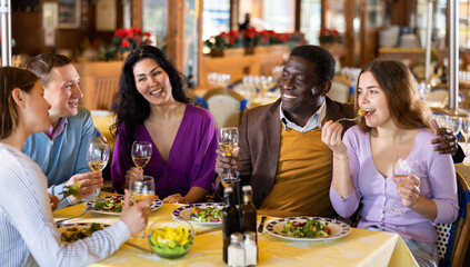 Wall Mural - Multiracial group of happy adult people having fun in cozy restaurant during dinner, chatting and drinking wine..