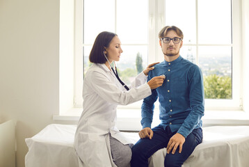 Female doctor with stethoscope listening to young man's heartbeat. Woman who works as physician checking male patient's lungs or heart sitting on examination couch in modern medical office exam room