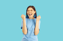 Beautiful Young Woman Wins Cool Prize Or Is Excited About Holiday Sale. Happy Euphoric Lady In Blue T Shirt And Earrings Standing On Blue Background, Fist Pumping And Screaming YES, I DID IT, HOORAY