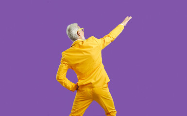 Wall Mural - Back view of funny energetic senior man in stylish outfit having fun at party. Backview of cheerful elderly guy wearing bright yellow party suit dancing isolated on solid purple colour background