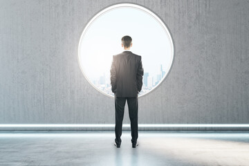 Poster - Back view of thoughtful young businessman standing in creative modern concrete interior with round window and city view. Mock up place on wall and daylight.