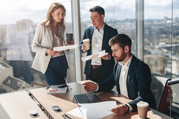 Wall Mural - Focused businessmen drink coffee and discussing documents with graphs and charts in a modern office.