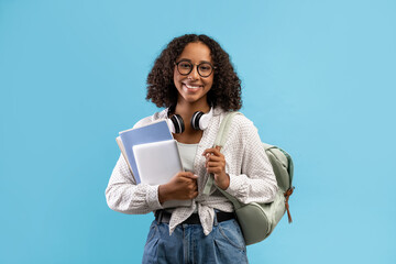 Online education. Smiling black female student with backpack and headphones holding notebooks and digital tablet