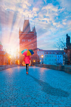 Woman In Red Clothes With Multicolored Umbrella - View Of Charles Bridge And Lesser Bridge Tower In Prague, Czech Republic