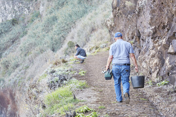 Rearview of a man walking on a narrow cliff carrying pale and toolbox