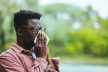 Allergic Black Man Blowing On Wipe In A Park On Spring Season. Man With Allergy Or Cold, Blowing His Nose With A Tissue, Looking Miserable Unwell Very Sick, Isolated Outside Green Trees Background. 