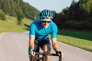 Wall Mural - Caucasian man in professional road cycling clothes, a blue jersey, with a helmet, sunglasses, and gloves, riding uphill on an asphalt road