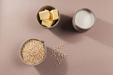 Wall Mural - Bowl of dry raw broken pearl barley cereal grain with butter and milk on beige background. Cooking pearl barley porridge concept.