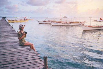 Wall Mural - Photography and travel. Young woman in hat holding camera sitting on wooden fishing pier with beautiful tropical sea view.