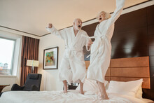 Senior Couple In White Robes Jumping On Bed And Looking Happy