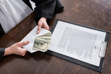Fototapeta  - cropped view of judge holding envelope with dollars near clipboard with lawsuit.