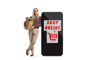 Poster - Full length portrait of a casual young woman with a grocery bag leaning on a smartphone with sign for online shopping