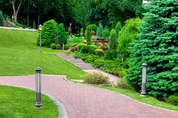 Wall Mural - iron lantern ground and pillar garden lighting of park curved path paved with stone tiles with drain lattice in park among plants, evergreen bushes and pine trees on hilly landscape, nobody.