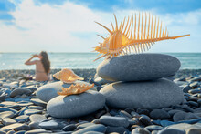 Sea, Ocean, Starfish, Shells, Girl Sitting On Shore Of Blue Sea In Rays Of Sunlight Against Background Of Blue Sky And Stones Of Pebble Shore. Concept Of Summer Holidays, Relaxation On Ocean.