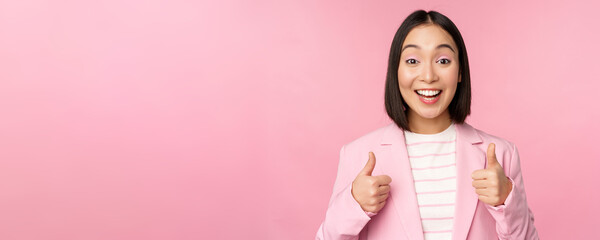 Wall Mural - Portrait of asian businesswoman smiling satisfied, showing thumbs up, praise, like and approve, standing in suit over pink background