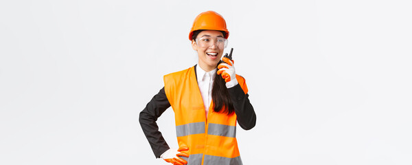 Wall Mural - Confident pleased asian female engineer in safety helmet and uniform talking with chief architect using walkie-talkie. Satisfied construction technician contact team using radio phone