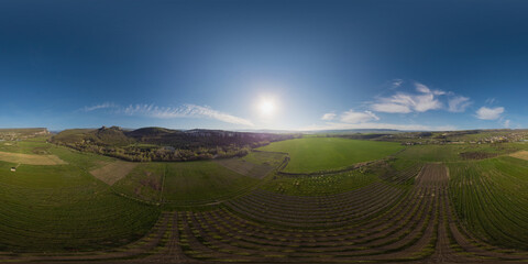  Aerial panorama over a young garden and a green wheat field in the countryside at sunset. Seamless spherical equirectangular 360 degree panorama. Agronomy, industry and food production.