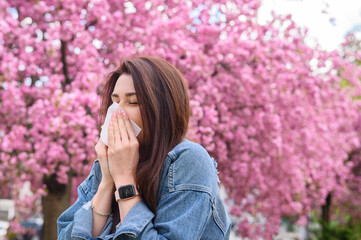 Young beautiful girl is allergic to flowers and blows her nose on a background of blooming pink sakura tree dressed in a blue denim jacket