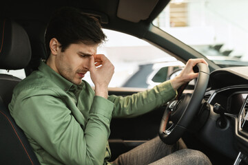 Unhappy young man sitting on driver's seat of new car, disappointed with test drive, having doubts about buying auto