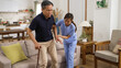 unwell asian elderly male with knee pain standing up slowly with a stick from the sofa at home. female nursing aide giving support while the man groans painfully