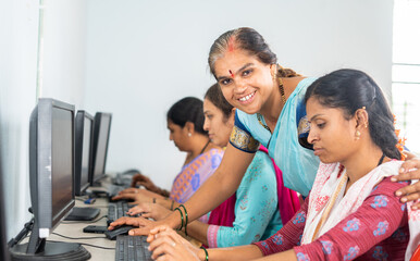 smiling teacher looking at camera while teaching computer class to woman's at village - concept of volunteer, self employment training and empowerment programs