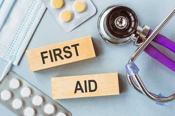 FIRST AID phrase on two wooden blocks on doctors table with stethoscope, white and yellow pills and medical face mask.