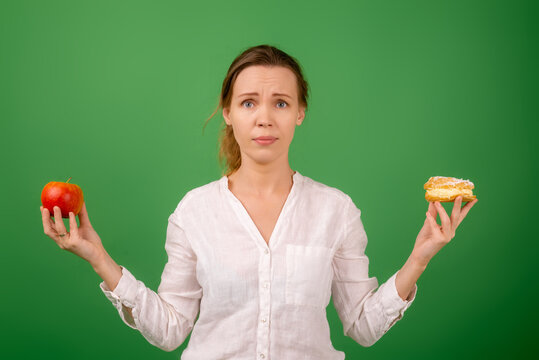 An attractive middle-aged woman in a white shirt holds an apple and a cake in each hand on a green background. The agony of choice. The concept of choosing the right healthy food.