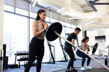 Fitness Class Together In A Gym Wearing Athletic Clothes Squatting While Holding A Barbell.