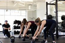 Three Woman In A Row Bent Over Doing Kettlebell Exercise While Personal Trainer Looks On In Background.