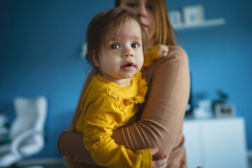 Wall Mural - One small caucasian baby girl happy with in hands of her mother holding her at home in front of blue wall in day real people front view copy space family bonding leisure concept