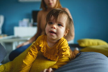 Wall Mural - One small caucasian baby girl happy with in hands of her mother holding her at home in front of blue wall in day real people front view copy space family bonding leisure concept