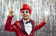 Lets get party. Positive funny young dark-skinned showman in shiny suit on silver foil background. Joyful crazy man in glasses, red hat and in suit with sequins raises his finger in sign of idea.