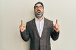 © Krakenimages.com - Middle age man with beard and grey hair wearing business jacket and glasses amazed and surprised looking up and pointing with fingers and raised arms.