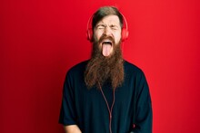 Redhead Man With Long Beard Listening To Music Using Headphones Sticking Tongue Out Happy With Funny Expression. Emotion Concept.
