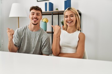 Poster - Young beautiful couple wearing casual clothes sitting on the table at home smiling with happy face looking and pointing to the side with thumb up.