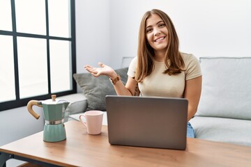 Sticker - Young brunette woman using laptop at home drinking a cup of coffee smiling cheerful presenting and pointing with palm of hand looking at the camera.