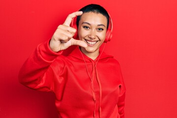 Poster - Beautiful hispanic woman with short hair listening to music using headphones smiling and confident gesturing with hand doing small size sign with fingers looking and the camera. measure concept.