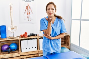 Poster - Middle age hispanic physiotherapist woman working at pain recovery clinic looking at the camera blowing a kiss with hand on air being lovely and sexy. love expression.