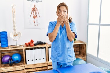 Canvas Print - Middle age hispanic physiotherapist woman working at pain recovery clinic shocked covering mouth with hands for mistake. secret concept.