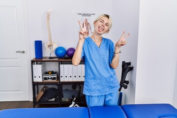 Sticker - Young physiotherapist woman working at pain recovery clinic smiling with tongue out showing fingers of both hands doing victory sign. number two.