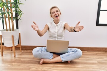 Young blonde woman using computer laptop sitting on the floor at the living room looking at the camera smiling with open arms for hug. cheerful expression embracing happiness.