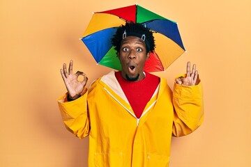 Poster - Young african american man wearing yellow raincoat looking surprised and shocked doing ok approval symbol with fingers. crazy expression