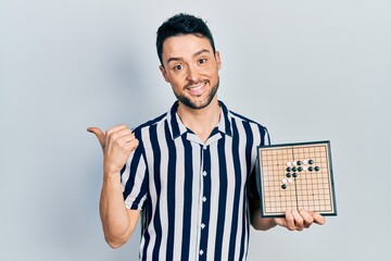 Poster - Young hispanic man holding asian go game board pointing thumb up to the side smiling happy with open mouth