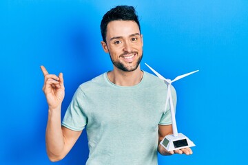 Young hispanic man holding solar windmill for renewable electricity smiling happy pointing with hand and finger to the side