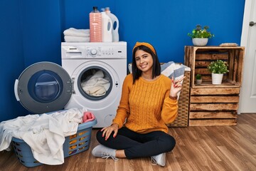 Poster - Young hispanic woman doing laundry with a big smile on face, pointing with hand finger to the side looking at the camera.