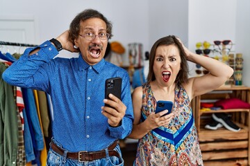 Poster - Middle age interracial couple at retail shop using smartphone crazy and scared with hands on head, afraid and surprised of shock with open mouth